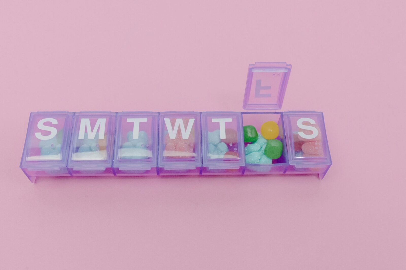 Colorful pills in a weekly organizer against a bright pink backdrop, representing daily medication routine.